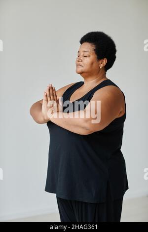 Portrait vertical d'une femme âgée noire méditant avec les yeux fermés tout en faisant du yoga à l'intérieur Banque D'Images