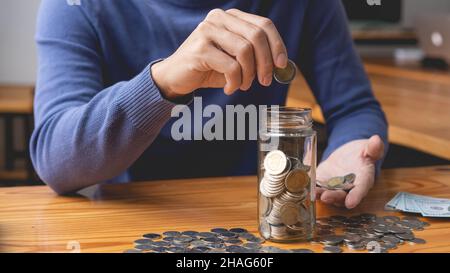 concept de sauvegarde l'homme dans le chandail bleu essayant d'économiser son argent en déposant les pièces de dix baht dans la bouteille. Banque D'Images
