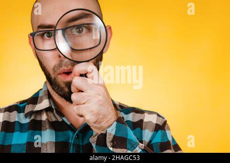 Le concept de la recherche et de la résolution des problèmes. Un homme chauve avec une barbe et des lunettes, portant une chemise à carreaux bleus, regarde à travers une loupe. Œil Banque D'Images