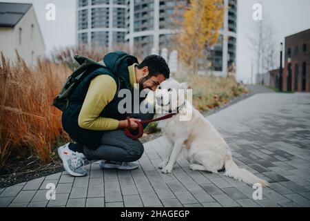 Un jeune homme heureux qui s'accroupette et embrasse son chien lors d'une promenade en plein air en ville. Banque D'Images