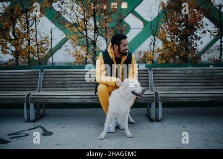 Jeune homme heureux assis sur le banc et tenant son chien à l'extérieur en ville en automne. Banque D'Images