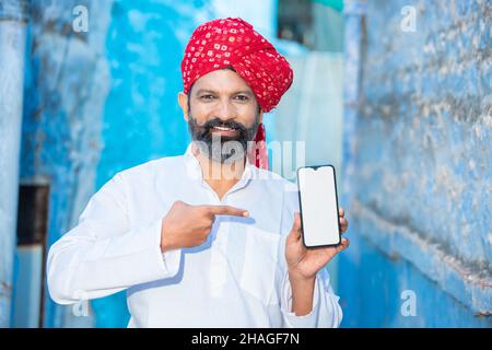 Un jeune homme indien traditionnel montrant une maquette d'écran de téléphone vierge, une personne portant une turban rouge avec une barbe affichant un smartphone vide pour la publicité d'un Banque D'Images