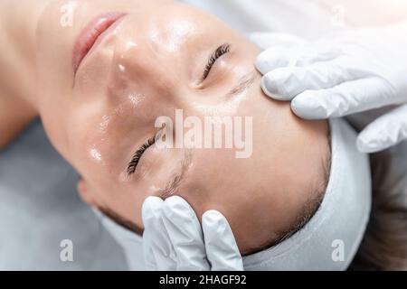 Gros plan esthéticienne médecin main fabrication anti-âge massage peeling pour jeune femme attrayante client à la clinique de beauté.Cosmétologue Banque D'Images