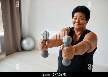 Portrait à la taille de la femme active senior levant des haltères tout en s'occupant à l'intérieur et en souriant à l'appareil photo Banque D'Images