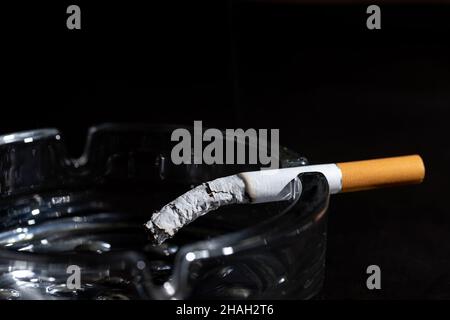 Gros plan la cigarette allumée avec belle wisp de fumée dans le cendrier avec beaucoup de mégots de cigarettes dans le plein et sale cendrier isolé sur fond noir co Banque D'Images