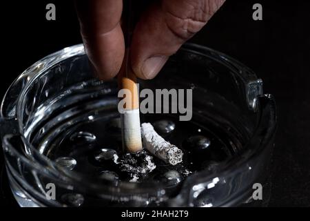 Gros plan la cigarette allumée avec belle wisp de fumée dans le cendrier avec beaucoup de mégots de cigarettes dans le plein et sale cendrier isolé sur fond noir co Banque D'Images