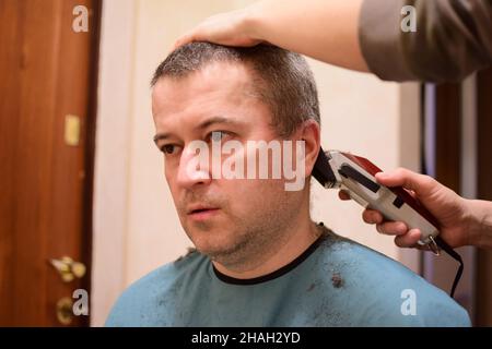 Un homme adulte est tondu avec une tondeuse à cheveux et s'assoit face à l'appareil photo.Peut-être à la maison dans un verrouillage Banque D'Images