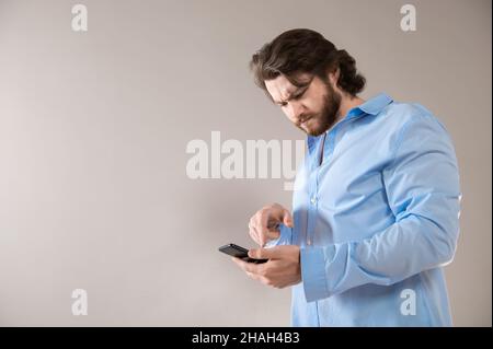 Homme barbu sérieux portant une chemise bleue avec smartphone isolé sur fond gris Banque D'Images