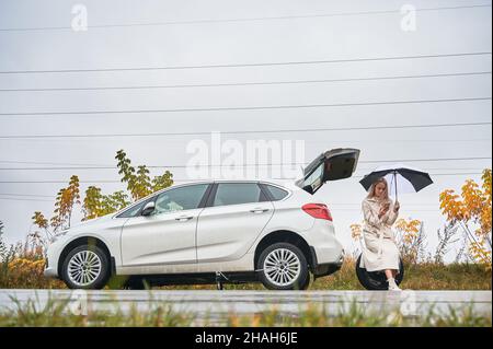 Belle femme sur la route près de son automobile blanche avec pneu de voiture crevé.Femme chauffeur appelant le service de voiture pour obtenir de l'aide, tenant un parapluie, assis sur la roue de secours. Banque D'Images