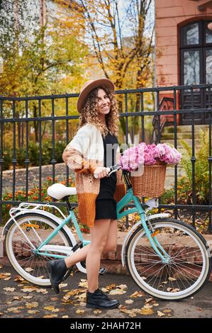 Vue latérale d'une femme maurique voyageant en vélo d'époque dans la rue.Belle femme portant chapeau brun et gilet est vélo d'équitation avec grand panier de camomille rose.Concept de loisirs. Banque D'Images