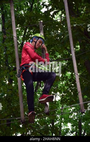 Un adolescent dans un casque de sécurité monte une échelle suspendue dans un parc d'attractions de corde Banque D'Images