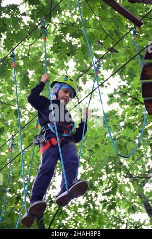 Un adolescent dans un casque de sécurité monte une échelle suspendue dans un parc d'attractions de corde Banque D'Images