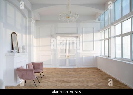 Intérieur d'un hall blanc spacieux avec fauteuils placés près d'une cheminée artificielle avec statuette et miroir sur lui en face de grandes fenêtres et grand chandeli Banque D'Images