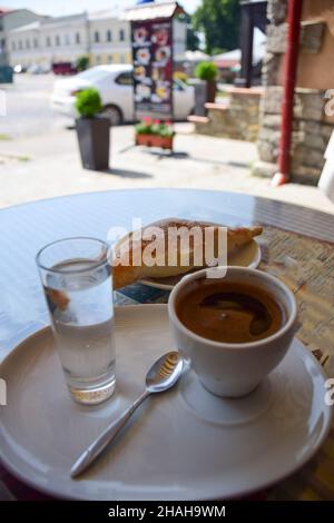 Une tasse de café, un croissant et un verre d'eau sur une soucoupe sont sur une table dans un café en bord de route.L'arrière-plan est très flou Banque D'Images