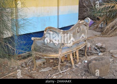 Un vieux canapé en ruines et déchiré avec ses côtés tombant dehors se trouve dans un jardin près d'une pile de déchets et d'un mur peint Banque D'Images