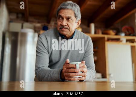 Pensive retraité biracial homme âgé se penchant sur le comptoir de cuisine moderne, tenant le café du matin dans ses mains. Banque D'Images