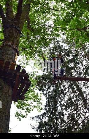 Un adolescent dans un casque de sécurité monte une échelle suspendue dans un parc d'attractions de corde Banque D'Images