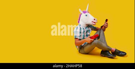 un homme portant un masque unicorn tient un téléphone fixe rouge coloré dans ses mains, assis sur un fond jaune, dans un format panoramique à utiliser comme web b Banque D'Images