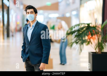 Portrait d'un jeune homme confiant dans un costume élégant portant un masque de protection face regardant loin dans le hall du centre commercial, arrière-plan flou. Banque D'Images