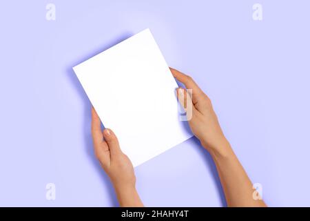 Vue de dessus des mains de femme tenant une carte de vœux ou d'invitation en papier vierge maquette sur fond pastel lilas.Couleur de l'année 2022.Noël, Saint-Valentin Banque D'Images