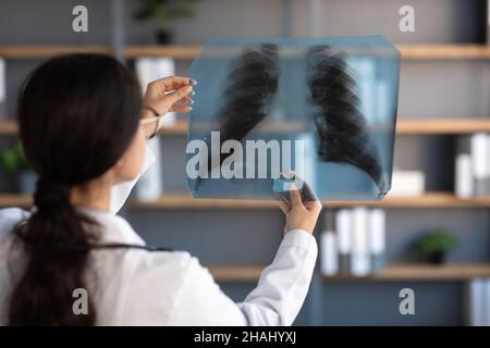 Jeune femme indienne médecin en manteau blanc regarde la photo de rayons X des poumons dans l'intérieur de clinique de bureau, dos Banque D'Images