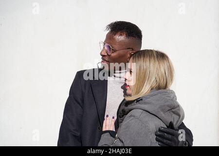 Homme noir en lunettes et manteau embrassant sa petite amie blonde en manteau par temps ensoleillé. Banque D'Images