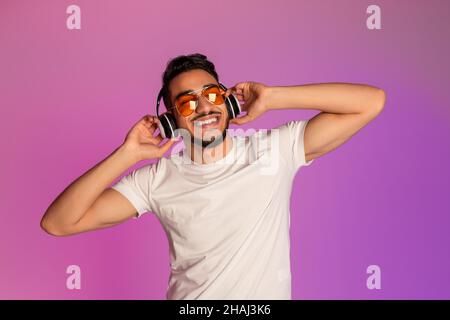 Jeune homme arabe décontracté à l'écoute de musique dans un casque sans fil, portant des lunettes de soleil, fermant les yeux dans la lumière du néon Banque D'Images