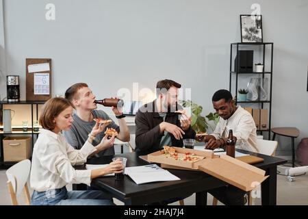 Groupe de collègues d'affaires qui boivent de la bière et mangent de la pizza tout en célébrant quelque chose à la table au bureau Banque D'Images