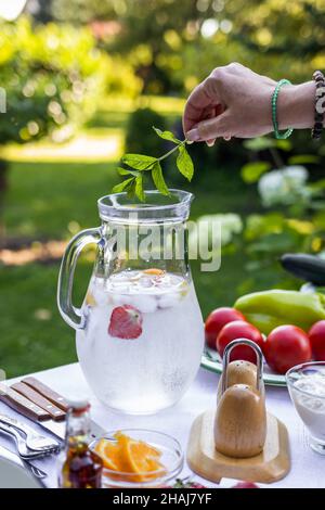 Femme à la main mettant l'herbe à la menthe dans la limonade froide dans le pichet.Préparation d'une boisson non alcoolisée pour la fête de jardin Banque D'Images