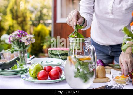 Femme mettant l'herbe de menthe dans la limonade froide dans le pichet.Préparation d'une boisson non alcoolisée pour la fête de jardin Banque D'Images