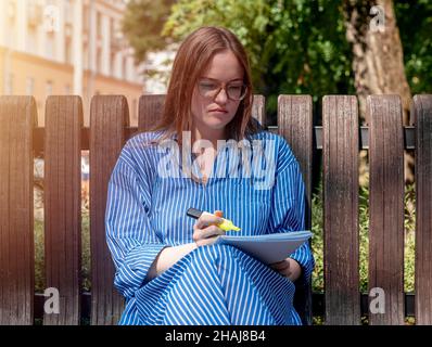 Étudiant en lunettes assis sur un banc dans le parc public de la ville avec syllabus ou bloc-notes avec surligneur.Femme étudiant à l'extérieur. Banque D'Images