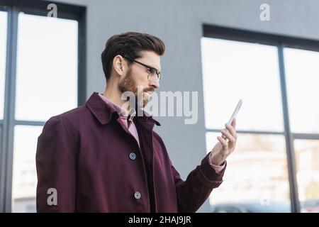 Homme d'affaires barbu en trench-coat utilisant un smartphone dans la rue urbaine Banque D'Images