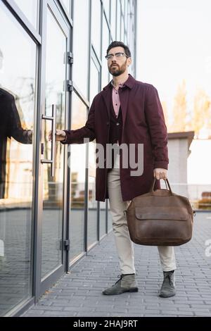 Homme d'affaires en trench coat tenant un sac près de la porte du bâtiment à l'extérieur Banque D'Images