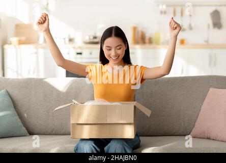 Service de livraison.Une femme asiatique surjoyeuse qui défait la boîte de colis, se moque des poings, heureuse de recevoir le paquet Banque D'Images