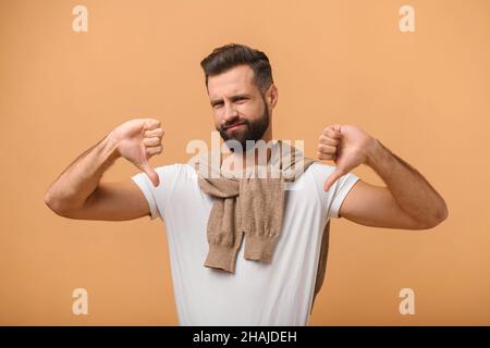 Portrait de l'homme avec la barbe montrant les pouces vers le bas, exprimant la désapprobation, critiquant et en désaccord avec la suggestion.Studio d'intérieur isolé sur fond orange Banque D'Images