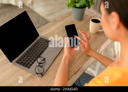 Jeune femme méconnaissable tenant un smartphone avec un écran vierge, assise à une table avec un ordinateur portable, maquette Banque D'Images