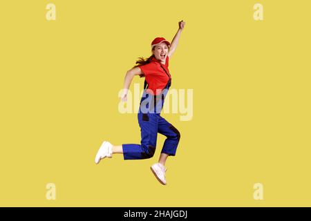 Portrait d'une femme très heureuse qui saute et se serre les poings, célébrant une journée de congé tant attendue, portant une combinaison et une casquette rouge.Studio d'intérieur isolé sur fond jaune. Banque D'Images