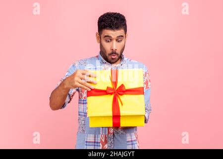 Portrait de curieux heureux homme cadeau d'ouverture, peeking à l'intérieur de la boîte avec le regard de nez, déballage du présent, en prévision d'une intéressante surprise d'anniversaire.Studio d'intérieur isolé sur fond rose. Banque D'Images