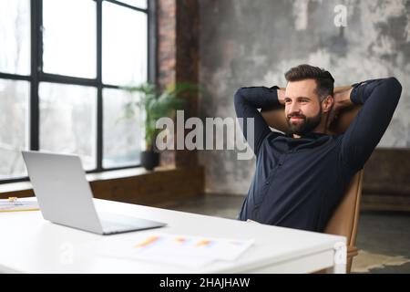 Homme sans rasé assis à la table et souriant, tout en se sentant à l'aise et à la détente sur son lieu de travail.Travailleur de bureau confiant assis devant l'ordinateur portable Banque D'Images
