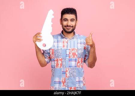 Portrait d'homme beau positif en chemise bleue tenant une énorme clé en papier et souriant satisfait de la nouvelle maison, montrant le pouce vers le haut, service de location.Studio d'intérieur isolé sur fond rose. Banque D'Images