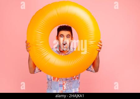 Homme barbu choqué portant une chemise bleue décontracté tenant l'anneau en caoutchouc dans les mains et regardant la caméra avec l'expression surprise et la bouche ouverte.Studio d'intérieur isolé sur fond rose. Banque D'Images