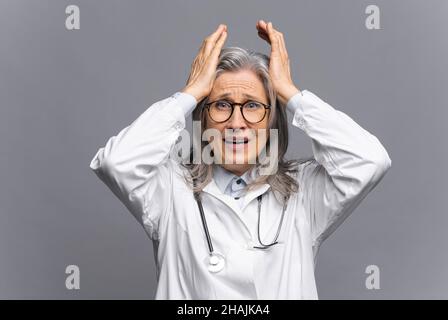 Médecin senior en robe médicale blanche avec stéthoscope sur les épaules regardant la caméra avec la peur sur le visage, la tête de préhension isolée sur le gris, thérapeute physique mature se sent désespéré et méprisant Banque D'Images