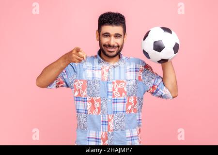 Jeune homme de barbe adulte avec un sourire crasseux portant une chemise bleue de style décontracté pointant sur vous, tenant le ballon de football, regardant l'appareil photo avec un look heureux.Studio d'intérieur isolé sur fond rose. Banque D'Images