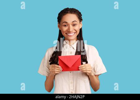 J'ai reçu la lettre d'amour le jour de la Saint-Valentin.Heureuse belle femme avec des dreadlocks noirs tenant la lettre dans l'enveloppe rouge ou carte de voeux et souriant joyeusement.Studio d'intérieur isolé sur fond bleu. Banque D'Images