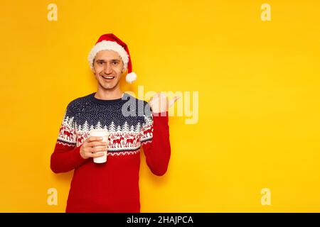 Un homme excité dans des vêtements de Noël chandail avec renne rouge chapeau de Père Noël tient le café de tasse de papier pour aller son doigt sur une zone vide pour la publicité isolée sur fond jaune avec espace pour le texte.Photo de haute qualité Banque D'Images