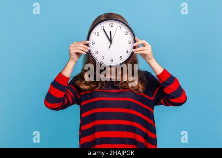 Portrait d'une femme anonyme inconnue portant un chandail rayé de style décontracté, cachant son visage, gestion du temps, horaire et rendez-vous de réunion.Studio d'intérieur isolé sur fond bleu. Banque D'Images