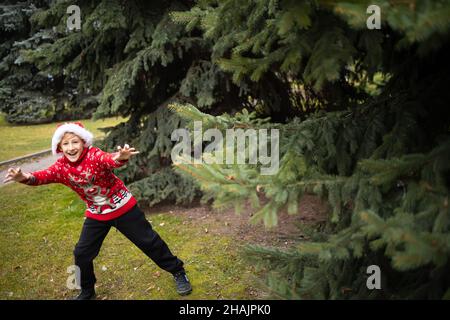 Un garçon dans un chandail de Noël tricoté rouge avec un renne de Noël et un chapeau de Père Noël saute de derrière les branches des arbres de Noël dans le Banque D'Images