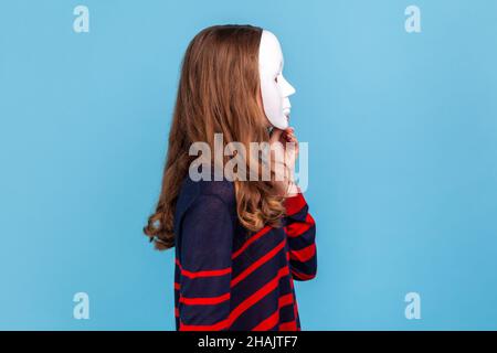 Vue latérale d'une femme inconnue portant un pull rayé de style décontracté, couvrant son visage avec un masque blanc, cachant sa personnalité, secret.Studio d'intérieur isolé sur fond bleu. Banque D'Images