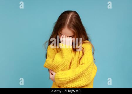 Portrait de petite fille se cachant face vers le bas et pleurant, bouleversé au sujet de la perte, sensation de tristesse et de regret, portant jaune style décontracté pull.Studio d'intérieur isolé sur fond bleu. Banque D'Images