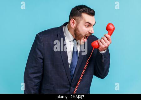 Un homme à barbe nerveux en colère portant un costume officiel hurlant et hurlant à parler d'un téléphone fixe rétro, se plaignant de la qualité de la connexion.Studio d'intérieur isolé sur fond bleu. Banque D'Images